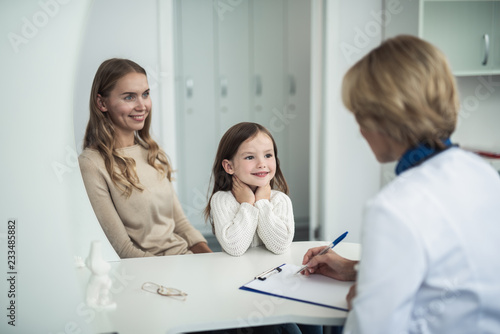 Concept of therapist consultation in healthcare system. Waist up back side portrait of pediatrician woman making notes while consulting mother and her daughter