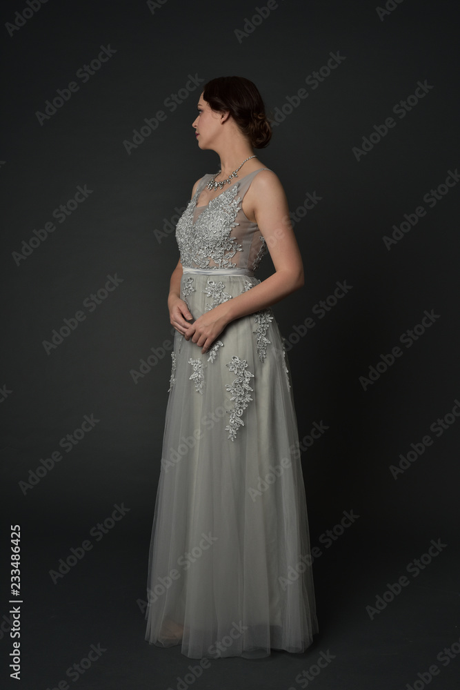 full length portrait of brunette  girl wearing long silver ball gown. standing pose on grey studio background.