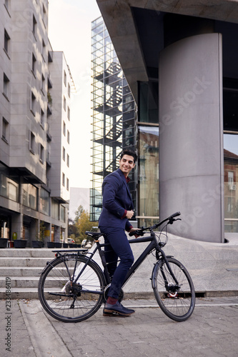 one young man, 20-29 years old, wearing suit, looking sideways, posing on fancy city bicycle. modern architecture building behind. full length shot.