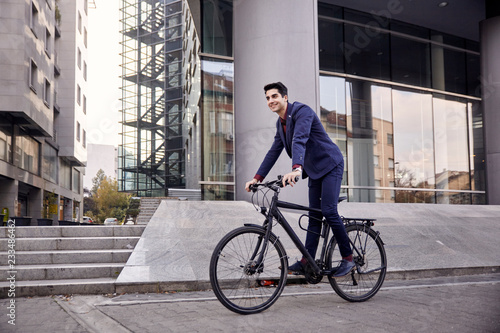 one young man, 20-29 years old, wearing suit, looking smiling. riding, pedaling standing, fancy bicycle. full length body. modern architecture building behind.
