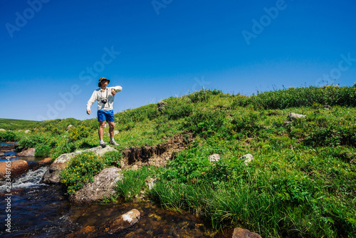 Traveler with camera on stone in mountain creek. Adventure of tourist. Hiking in mountains. Rich vegetation of highlands. Stream of clear water in brook. Vivid sunny landscape of majestic nature.