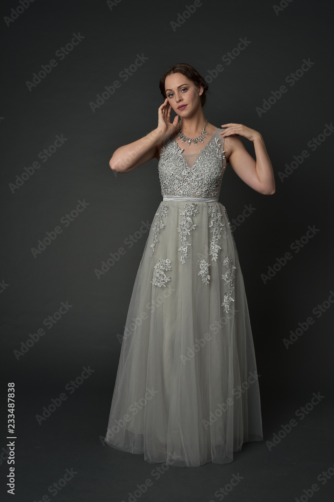 Fototapeta premium pełnej długości portret brunetka dziewczyna ubrana w długą srebrną suknię balową. poza stojącą na szarym tle studio.
