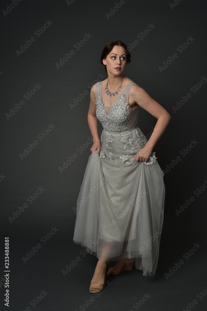 full length portrait of brunette  girl wearing long silver ball gown. standing pose on grey studio background.
