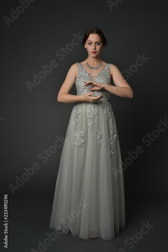 full length portrait of brunette girl wearing long silver ball gown. standing pose on grey studio background.