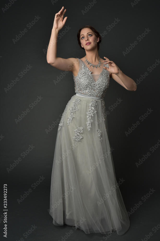 full length portrait of brunette  girl wearing long silver ball gown. standing pose on grey studio background.