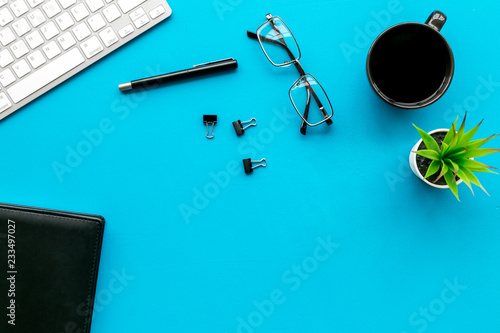 Office desk of creative person or hipster.Cute workplace. Keyboard and glasses near coffee, notebook, green room plant and stationery on blue, turquoise background top view copy space