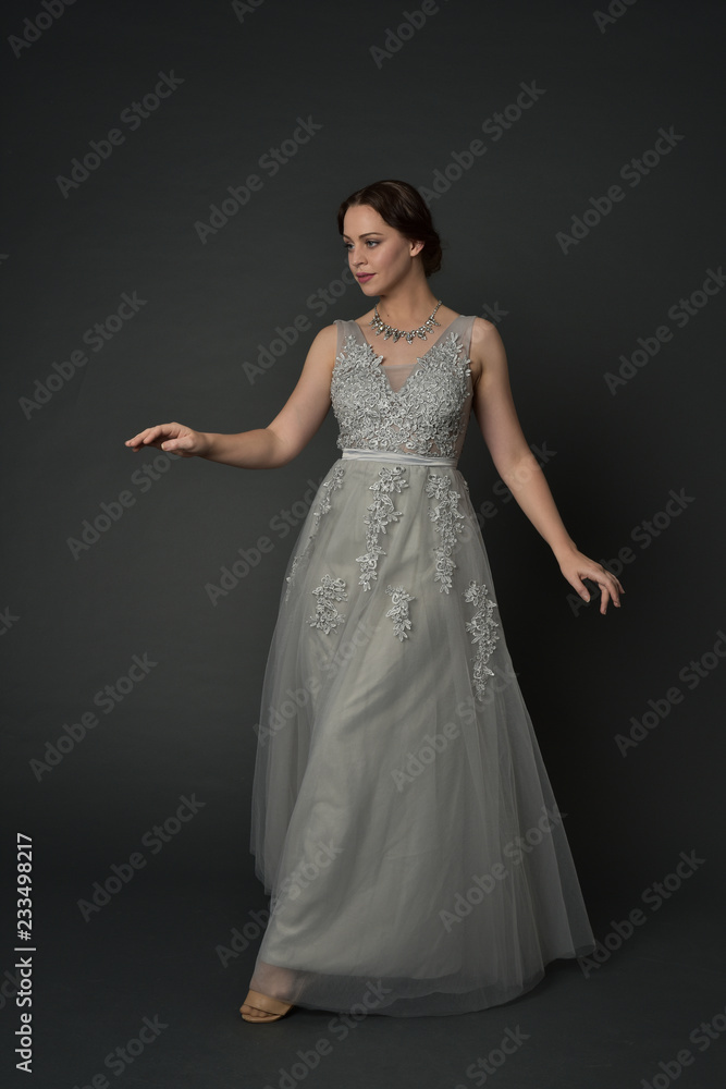full length portrait of brunette  girl wearing long silver ball gown. standing pose on grey studio background.