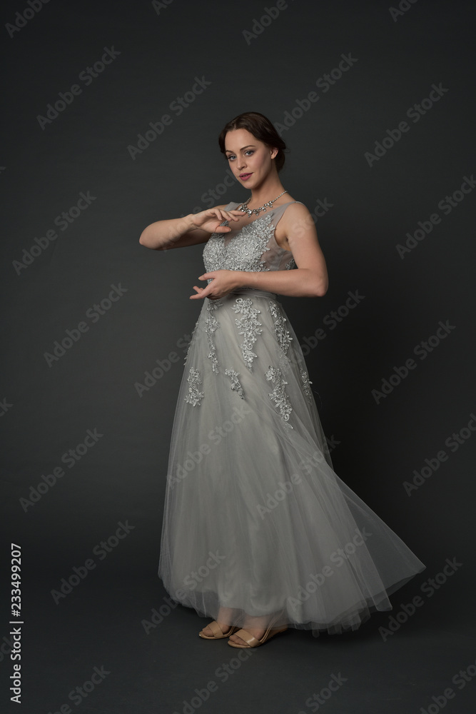 full length portrait of brunette  girl wearing long silver ball gown. standing pose on grey studio background.