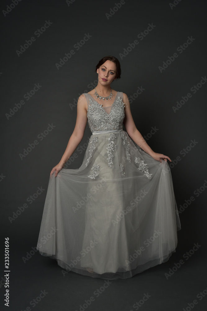 full length portrait of brunette  girl wearing long silver ball gown. standing pose on grey studio background.