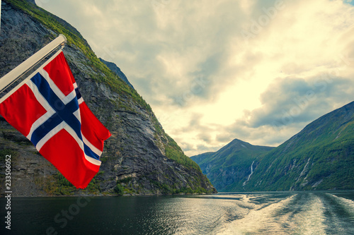 Mountain landscape with cloudy sky. Majestic Geiranger fjord.  View from ship. Norvegian flag against beautiful nature of Norway.