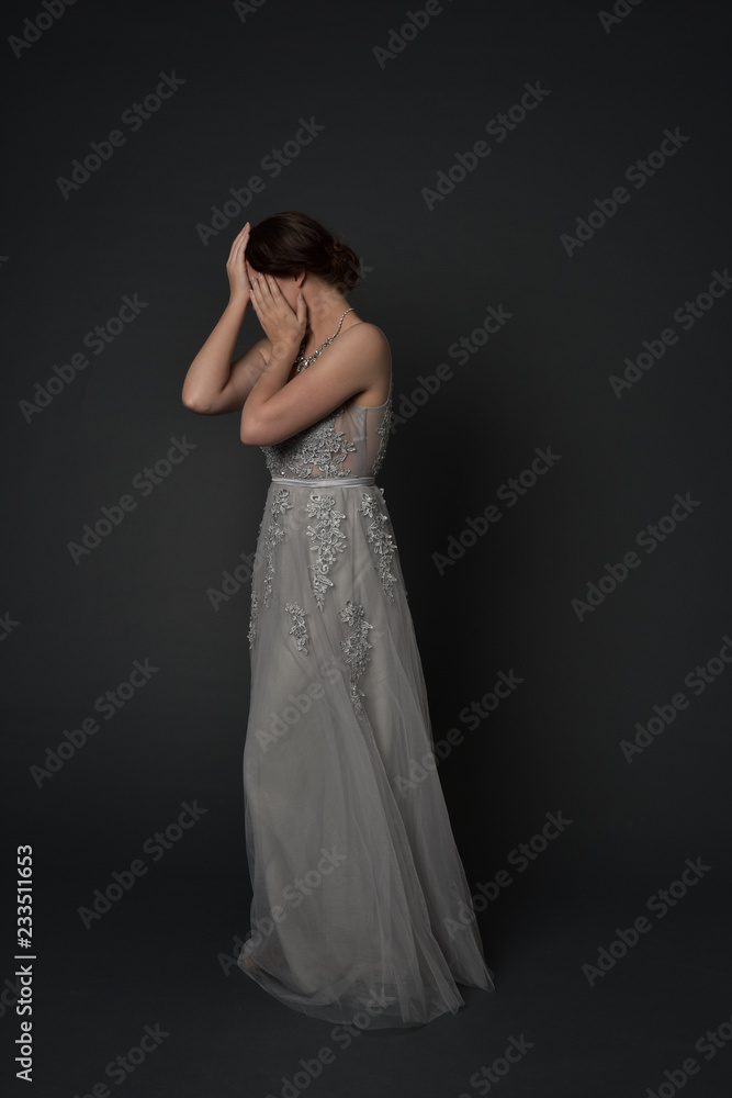full length portrait of brunette  girl wearing long silver ball gown. standing pose with hands covering face on grey studio background.