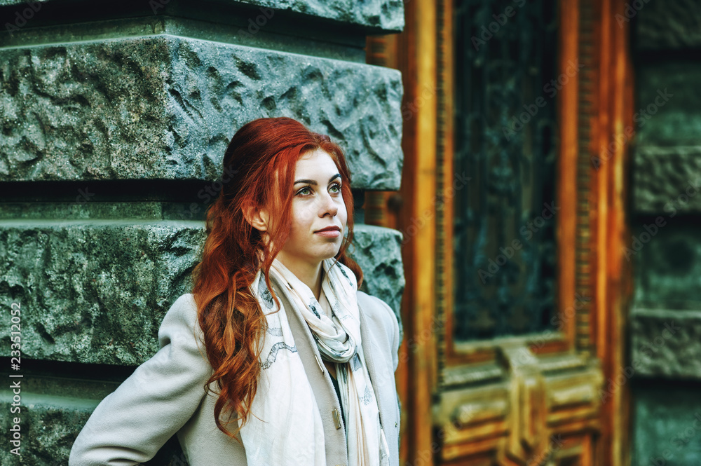 Fototapeta premium Portrait of a young beautiful fashionable red-haired woman with long curly hair , posing on the streets of the European city. The girl is dressed in a light coat