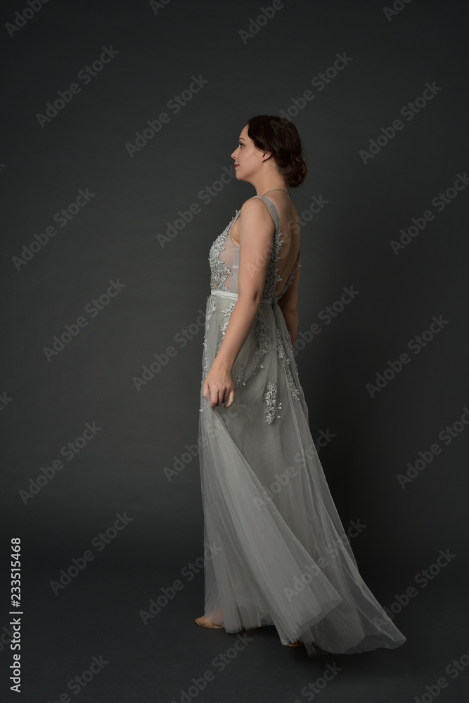 Fototapeta premium full length portrait of brunette girl wearing long silver ball gown. standing pose on grey studio background.