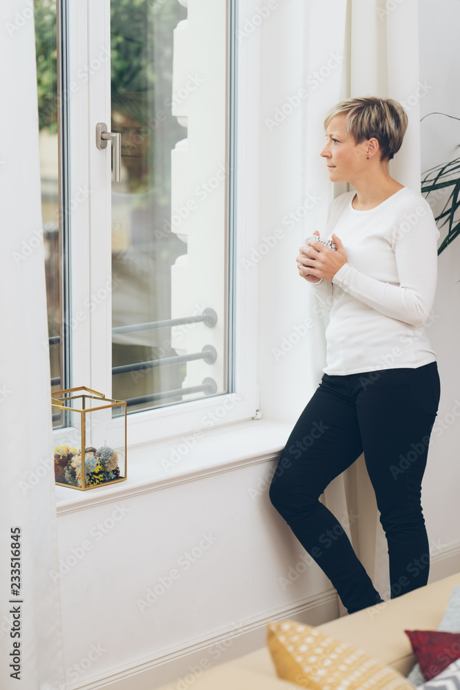 Woman standing looking out of a window at home Woman standing looking out of a window at home