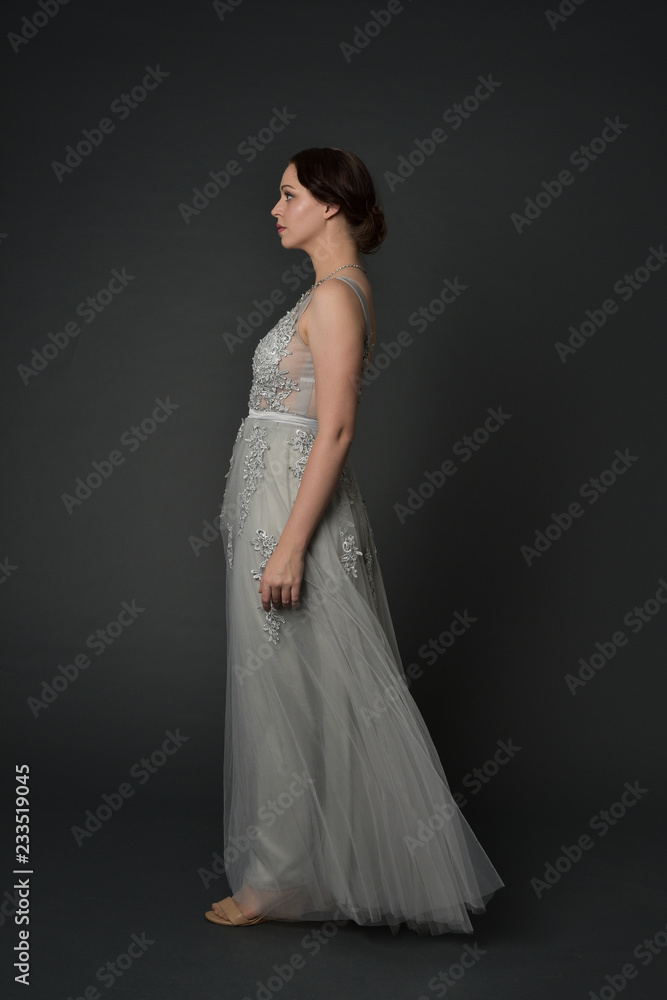 full length portrait of brunette girl wearing long silver ball gown. standing pose on grey studio background. full length portrait of brunette girl wearing long silver ball gown. standing pose on grey studio background.