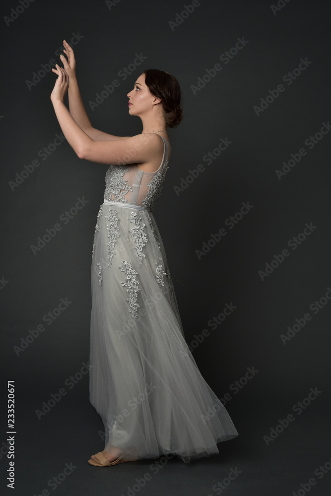Fototapeta premium full length portrait of brunette girl wearing long silver ball gown. standing pose on grey studio background.