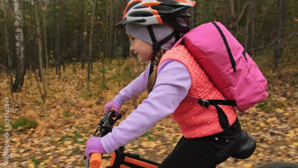 One caucasian children rides bike road in autumn park. Little girl riding black orange cycle in forest. Kid goes do bicycle sports. Biker motion ride with backpack and helmet. Mountain bike hardtail. One caucasian children rides bike road in autumn park. Little girl riding black orange cycle in forest. Kid goes do bicycle sports. Biker motion ride with backpack and helmet. Mountain bike hardtail.