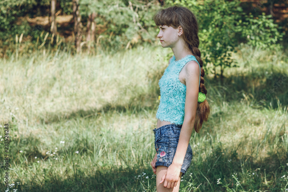 Young fashionable girl in denim shorts and a short top is standing against the background of green forest
