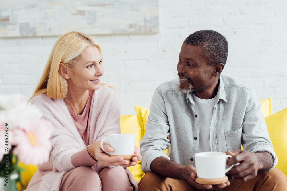 smiling mature woman sitting on sofa with african american man and drinking coffee together smiling mature woman sitting on sofa with african american man and drinking coffee together