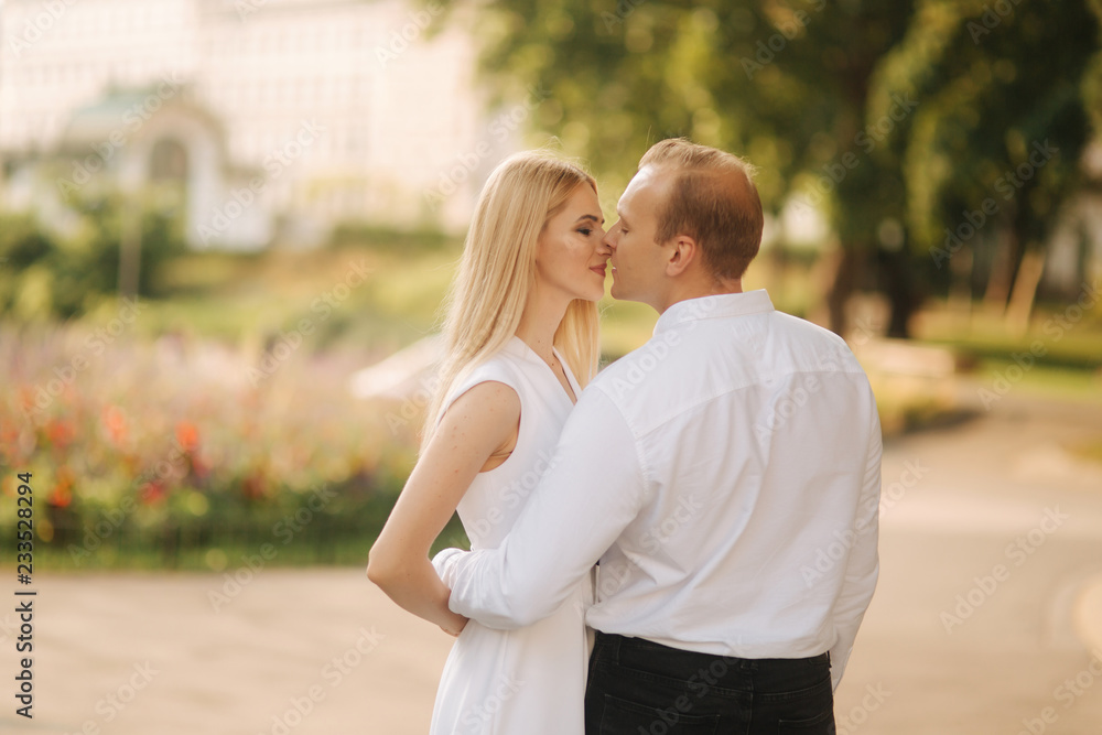Fototapeta premium Young beautiful couple husband in a white shirt and a woman in a dress walking around the park in summer time