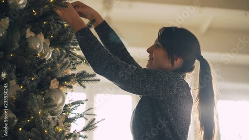 Beautiful young lady in sweater is decorating New Year tree with stylish decorations balls and lights hanging accessories on branches. People and events concept. photo