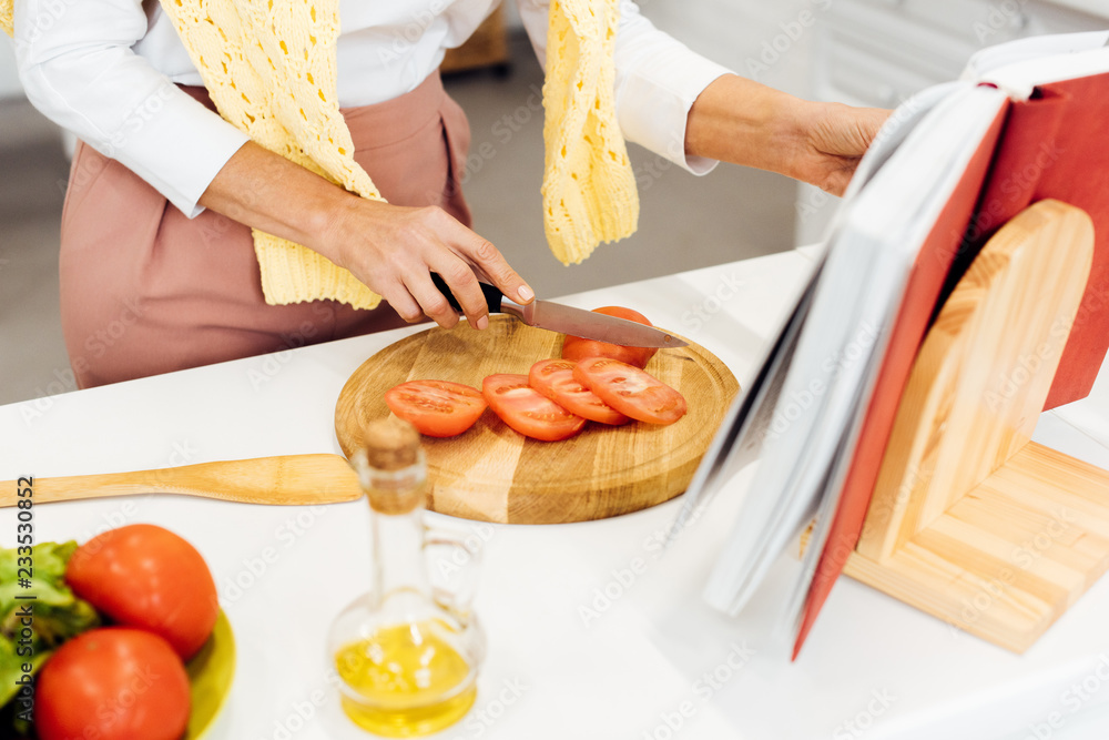 close up of woman slicing tomatoes and reading recipe in cookbook at kitchen