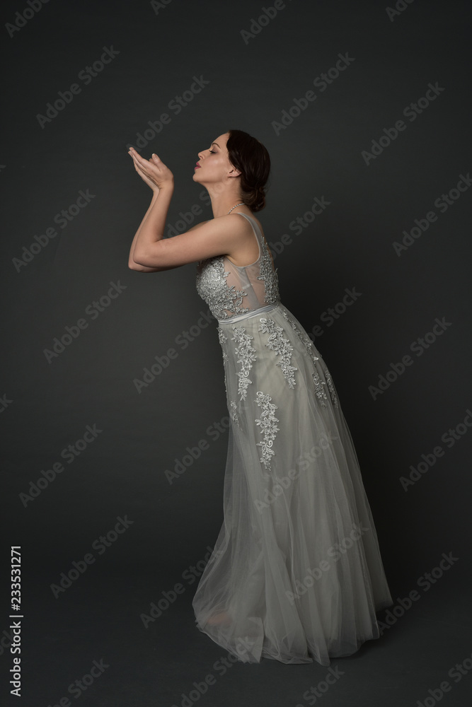 full length portrait of brunette  girl wearing long silver ball gown. standing pose on grey studio background.