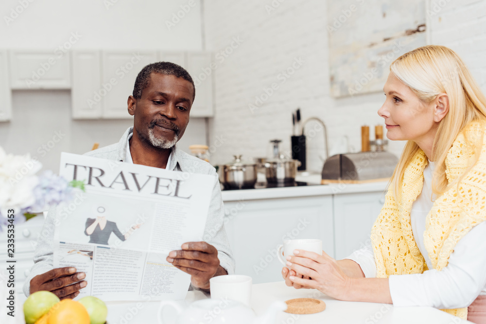 Obraz premium african american man reading newspaper while smiling mature woman drinking tea