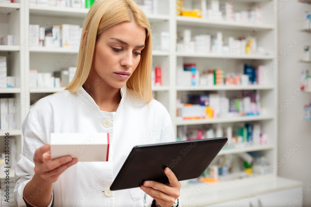 Serious young female pharmacist holding a tablet and a box of prescription drugs, standing in front of shelves full of medication boxes. Medicine, pharmaceutics, health care and people concept