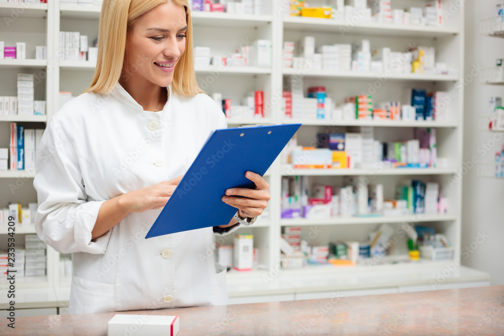 Happy young female pharmacist standing behind the counter with a clipboard, filling paperwork and writing. Medicine, pharmaceutics, health care and people concept Happy young female pharmacist standing behind the counter with a clipboard, filling paperwork and writing. Medicine, pharmaceutics, health care and people concept