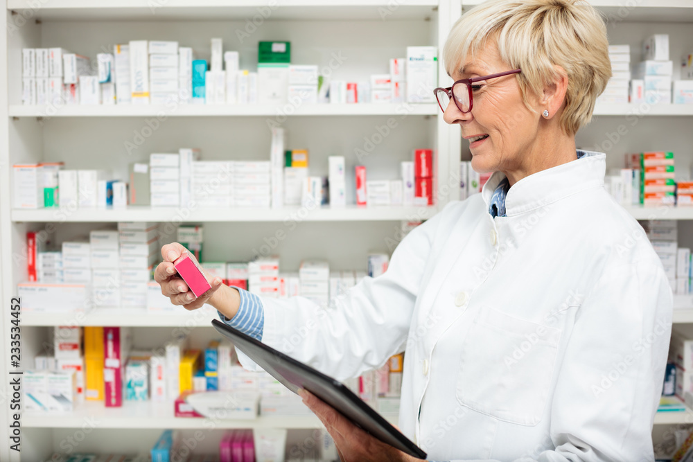 Happy mature female pharmacist holding a tablet and a box of prescription drugs. Shelf with medication boxes in the background. Medicine, pharmaceutics, health care and people concept