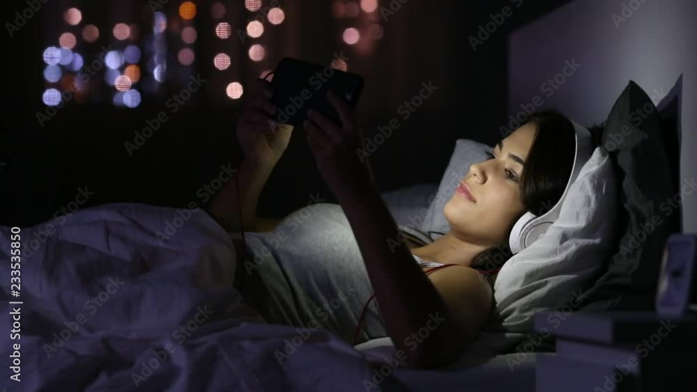 Happy teenage girl selecting and watching streaming video on a smart phone lying on the bed in the night Happy teenage girl selecting and watching streaming video on a smart phone lying on the bed in the night