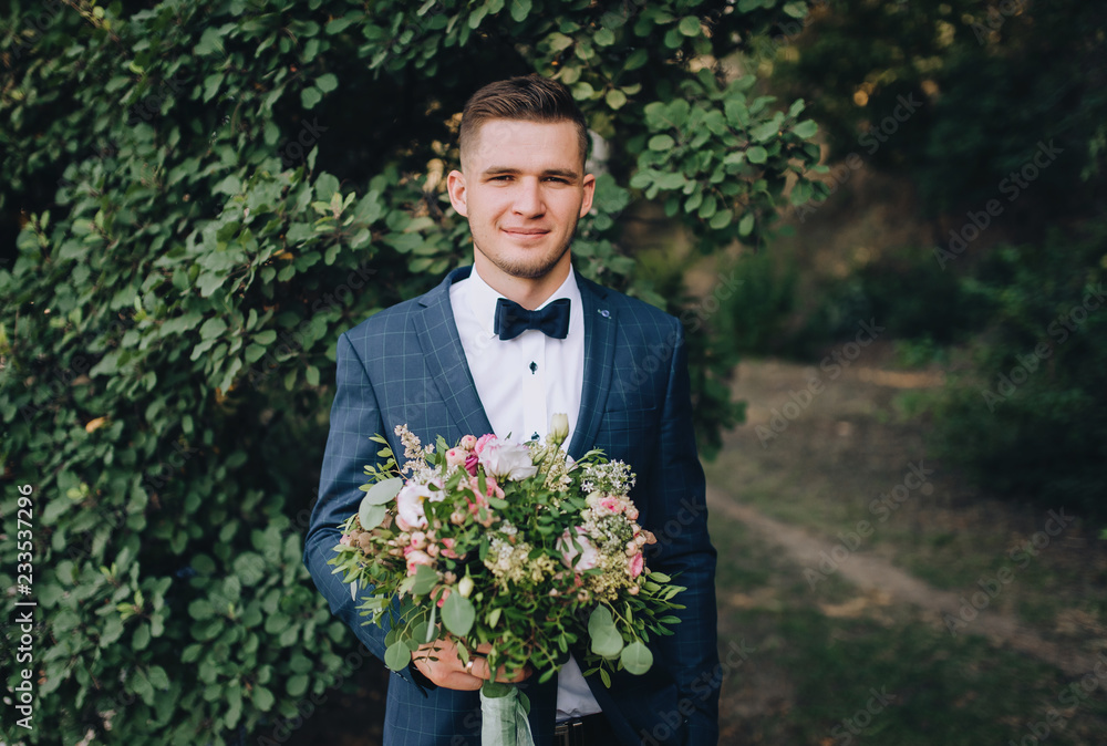 A young and stylish groom stands on the background of nature with green foliage, with a bouquet in his hands. Wedding portrait. Family photo. A young and stylish groom stands on the background of nature with green foliage, with a bouquet in his hands. Wedding portrait. Family photo.