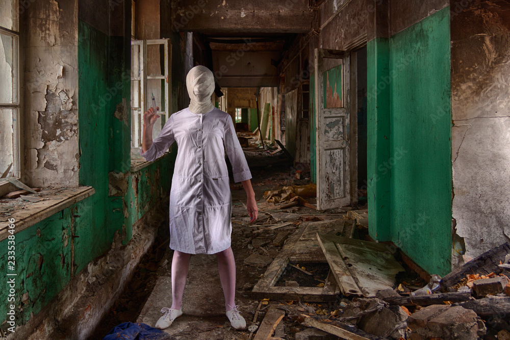 A ghost nurse or a zombie nurse or doctor with a tightly bandaged head and a face without eyes and mouth stands inside an abandoned clinic or asylum and holds a syringe in her hand. Horror scene
