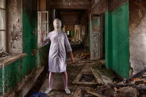 A ghost nurse or a zombie nurse or doctor with a tightly bandaged head and a face without eyes and mouth stands inside an abandoned clinic or asylum and holds a syringe in her hand. Horror scene © evgenzz
