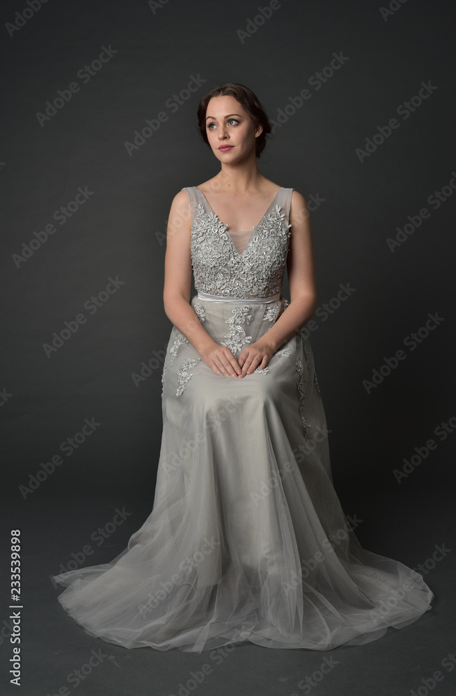 Fototapeta premium full length portrait of brunette girl wearing long silver ball gown. seated pose on grey studio background.