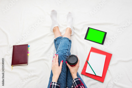 Girl in blue jeans and plaid shirt, holds in her hands coffee from disposable cup, tablet pc, women's feet, lying on white crumpled blanket. Background with copy space, for advertisement. Top view