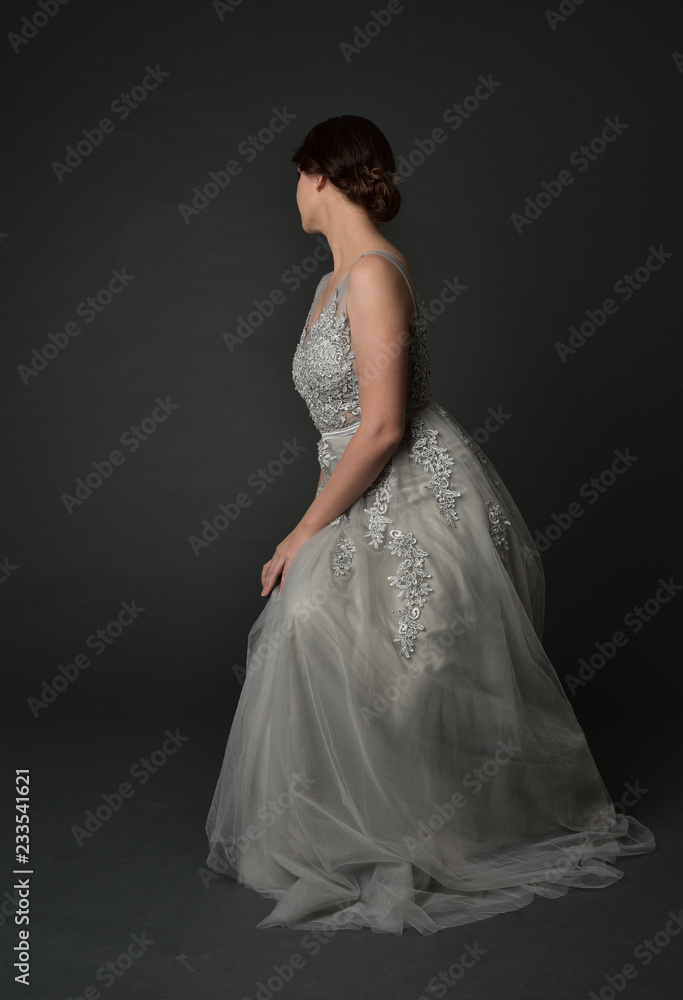  full length portrait of brunette  girl wearing long silver ball gown. seated pose on grey studio background.