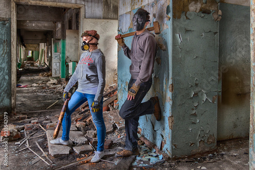 A pair of cyberpunk hooligans: a girl in gas mask and steampunk goggles with a bat and a guy in skull mask with a sledgehammer, standing in the middle of the ruins of an old building. street band