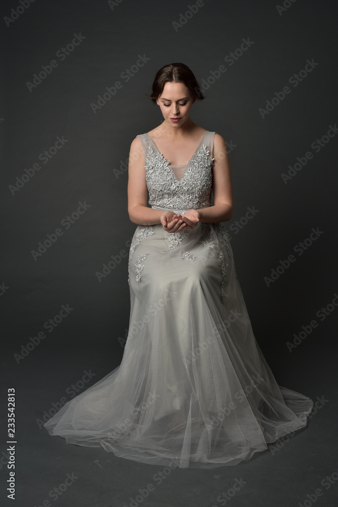 Fototapeta premium full length portrait of brunette girl wearing long silver ball gown. seated pose on grey studio background.