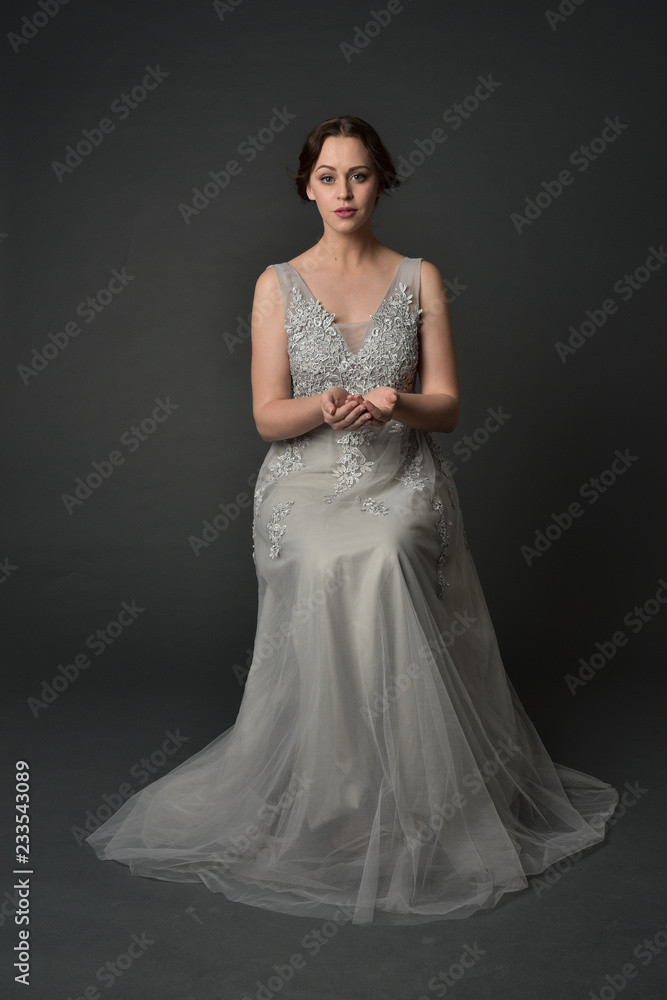 full length portrait of brunette girl wearing long silver ball gown. seated pose on grey studio background. full length portrait of brunette girl wearing long silver ball gown. seated pose on grey studio background.