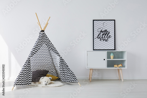Patterned black and white scandinavian tent with grey and yellow pillows and white teddy bear next to wooden cabinet with poster in black frame, copy space on empty white wall