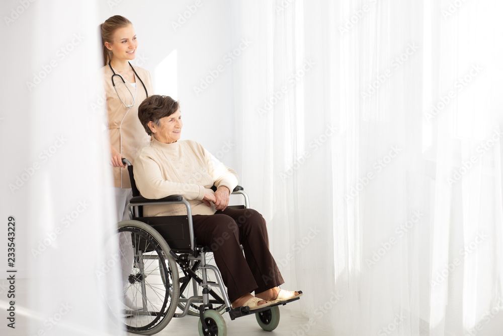 Smiling nurse supporting sick elderly woman in the wheelchair next to copy space Smiling nurse supporting sick elderly woman in the wheelchair next to copy space
