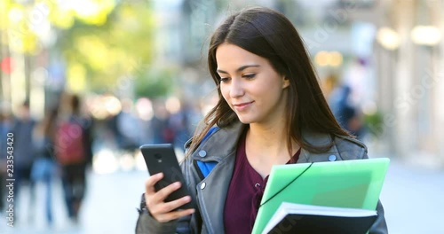 Happy student receiving a message checking smart phone stopped in the street after walking 