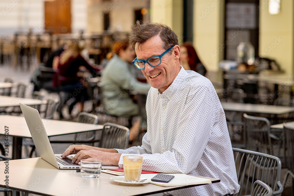 Happy attractive stylish mature man working on laptop while having coffee in outside coffee shop Happy attractive stylish mature man working on laptop while having coffee in outside coffee shop