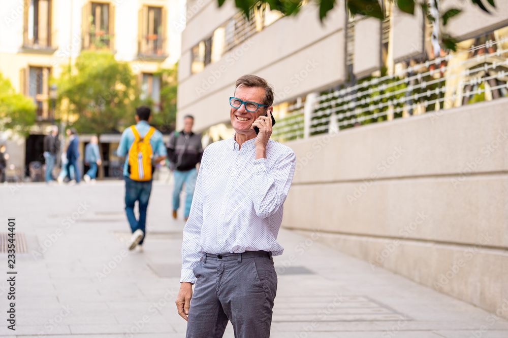 Happy attractive mature man on his smart phone while walking in the street smiling