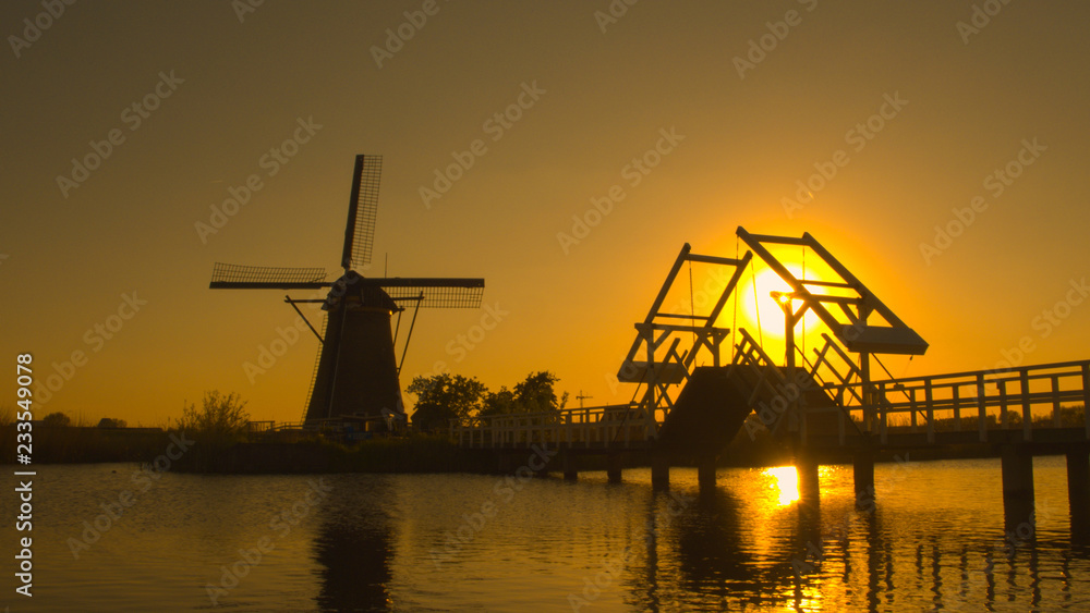 Wooden drawbridge on river with traditional windmill in background at sunset