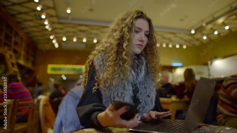 Young beautiful girl working on laptop in crowded restaurant, holding smartphone in other hand. Young beautiful girl working on laptop in crowded restaurant, holding smartphone in other hand.