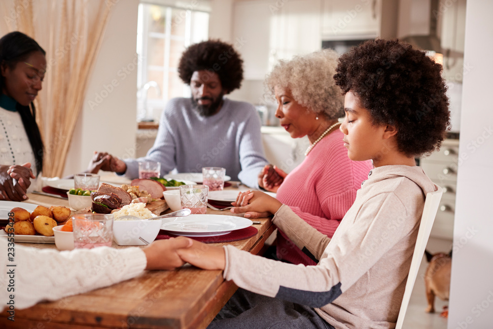 Multi generation mixed race family holding hands and saying grace before eating their Sunday dinner, side view Multi generation mixed race family holding hands and saying grace before eating their Sunday dinner, side view