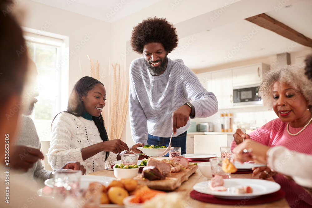 Middle aged black man carving and serving meat at Sunday family dinner with his partner, kids and their grandparents, front view Middle aged black man carving and serving meat at Sunday family dinner with his partner, kids and their grandparents, front view